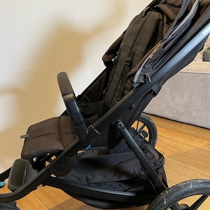 May include: A black stroller with a brown seat and a large storage basket. The stroller has a black canopy and a black handle.