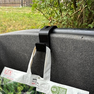 May include: A black plastic bag holder attached to a dark grey surface. A white shopping bag with a green forest print hangs from the holder. The bag has white handles and text.