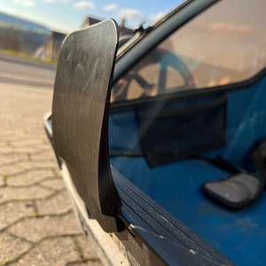Puede incluir: Primer plano de una cubierta de pl&aacute;stico negro para la ventana trasera de un veh&iacute;culo. La cubierta est&aacute; unida al marco de la ventana con una bisagra de metal negro.