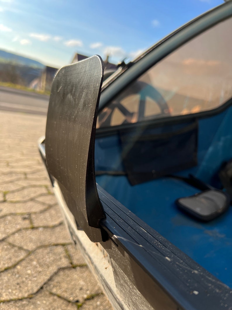 Puede incluir: Primer plano de una cubierta de pl&aacute;stico negro para la ventana trasera de un veh&iacute;culo. La cubierta est&aacute; unida al marco de la ventana con una bisagra de metal negro.
