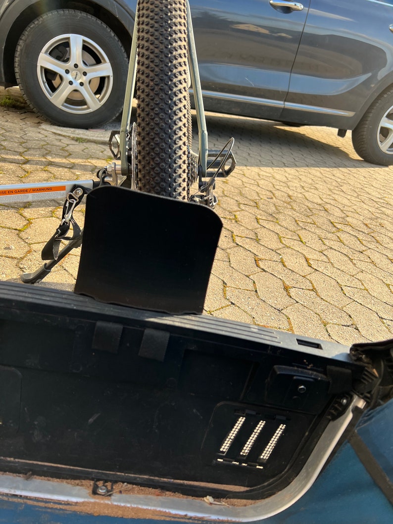 Puede incluir: Un portabicicletas negro fijado a la parte trasera de un coche plateado. El portabicicletas est&aacute; vac&iacute;o y tiene una solapa de metal negro que est&aacute; parcialmente abierta. El coche est&aacute; aparcado en una entrada de adoquines grises.