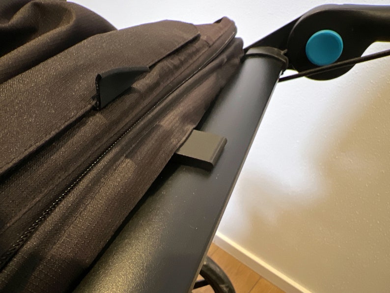 May include: Close-up of a black stroller canopy with a zipper and a black plastic clip. The canopy is attached to a black stroller frame with a blue button.