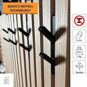 Puede incluir: Ganchos de metal negro montados en una pared de madera. Los ganchos están diseñados para instalarse rápida y fácilmente. El texto "QUICK 2 INSTALL TECHNOLOGY" es visible en la parte superior de la imagen. Los ganchos pueden soportar hasta 7 kg.