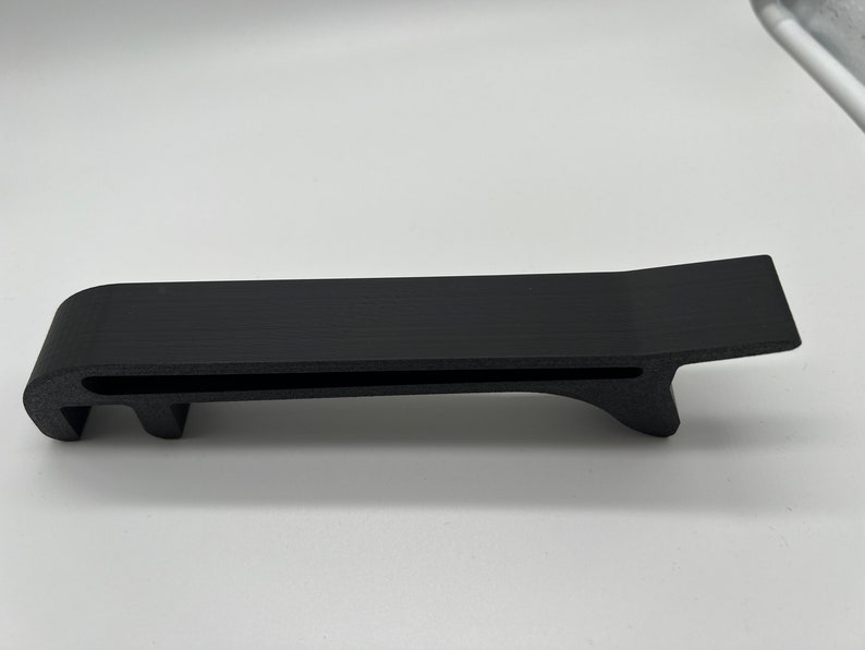 May include: A black plastic 3D printed object with a flat top and a curved bottom. The object has a slot cut into the top and two prongs on the bottom.