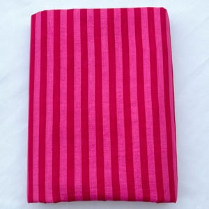 Könnte beinhalten: Ein rechteckiges Stück Stoff mit einem lebendigen Muster aus abwechselnden vertikalen Streifen in den Farben Pink und Magenta. Das Textil hat eine glatte, gleichmäßige Textur und ist zu einer sauberen, flachen, rechteckigen Form gefaltet.