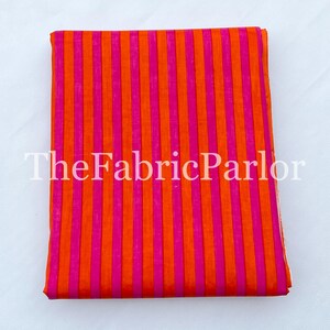 Puede incluir: Una pieza de tela doblada que presenta un vibrante patr&oacute;n de rayas verticales en colores naranja brillante y rosa intenso alternados. El texto TheFabricParlor est&aacute; superpuesto en blanco sobre el centro de la tela, que est&aacute; apilada.