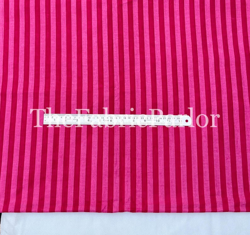 Puede incluir: Una tela con rayas verticales alternas en dos tonos de rosa. Una regla met&aacute;lica est&aacute; colocada horizontalmente en el centro, mostrando medidas en pulgadas y cent&iacute;metros, con el texto The Fabric Parlor superpuesto.