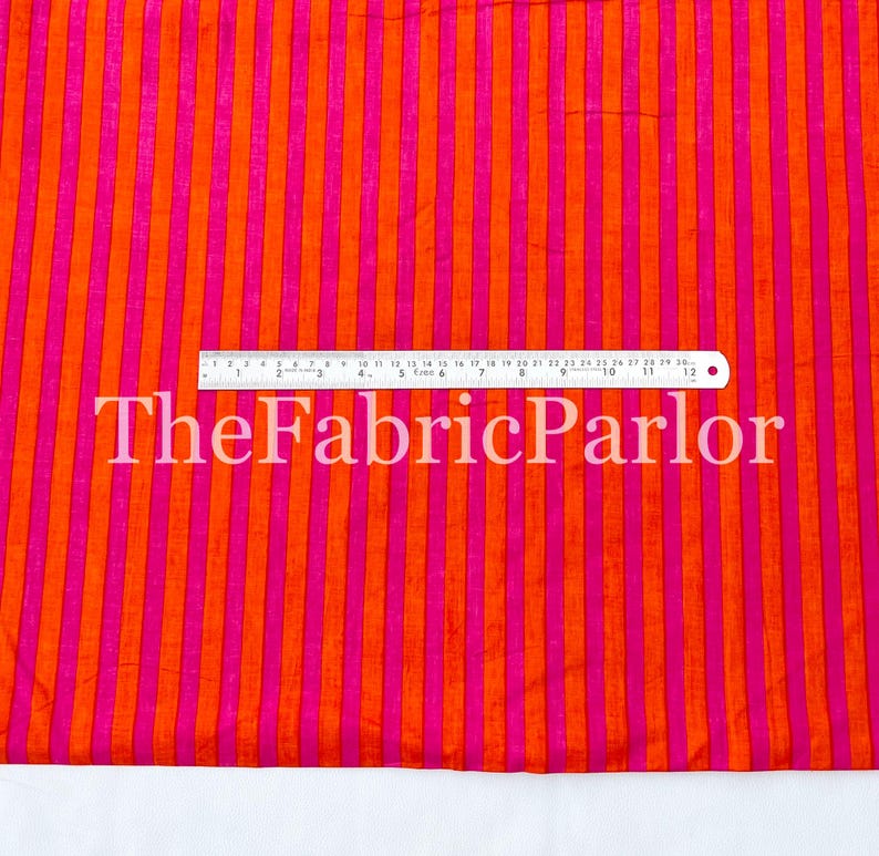 Puede incluir: Una tela con rayas verticales que alternan entre naranja brillante y rosa intenso. Una regla est&aacute; colocada horizontalmente en el centro, mostrando medidas de hasta 30 cm. El texto TheFabricParlor est&aacute; superpuesto en blanco en el medio.