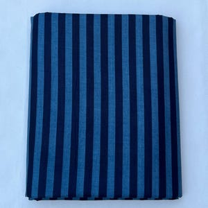 May include: A rectangular piece of fabric featuring a repeating vertical stripe pattern in alternating shades of dark navy blue and medium denim blue. The textile is folded into a neat, flat stack with clean, straight edges and a consistent weave.