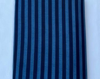 Puede incluir: Una pieza de tela rectangular que presenta un patrón de rayas verticales repetitivas en tonos alternos de azul marino oscuro y azul mezclilla medio. El tejido está doblado en una pila plana y ordenada con bordes rectos y consistentes.
