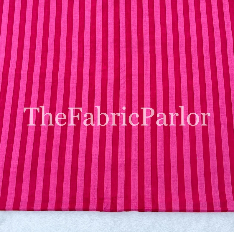 Puede incluir: Una tela textil con un patr&oacute;n repetitivo de rayas verticales en tonos alternos de rosa brillante y magenta intenso. El texto TheFabricParlor est&aacute; superpuesto en blanco en el centro de la superficie de la tela a rayas.
