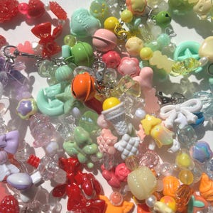 Puede incluir: Una colección de abalorios y cuentas coloridas y caprichosas de varias formas y tamaños. El surtido incluye corazones, estrellas, lazos, conos de helado y otros elementos decorativos en tonos de rojo, rosa, verde, amarillo y transparente. Algunos abalorios tienen el texto "HOT LOVE".