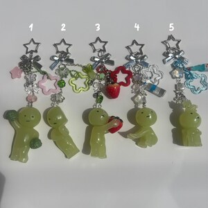 Peut inclure: Cinq porte-cl&eacute;s avec des fermoirs en forme d'&eacute;toile et divers breloques. Chaque porte-cl&eacute;s pr&eacute;sente une figurine verte et des &eacute;l&eacute;ments d&eacute;coratifs comme des &eacute;toiles, des fleurs et une fraise. Les porte-cl&eacute;s sont dispos&eacute;s en rang&eacute;e sur fond blanc.