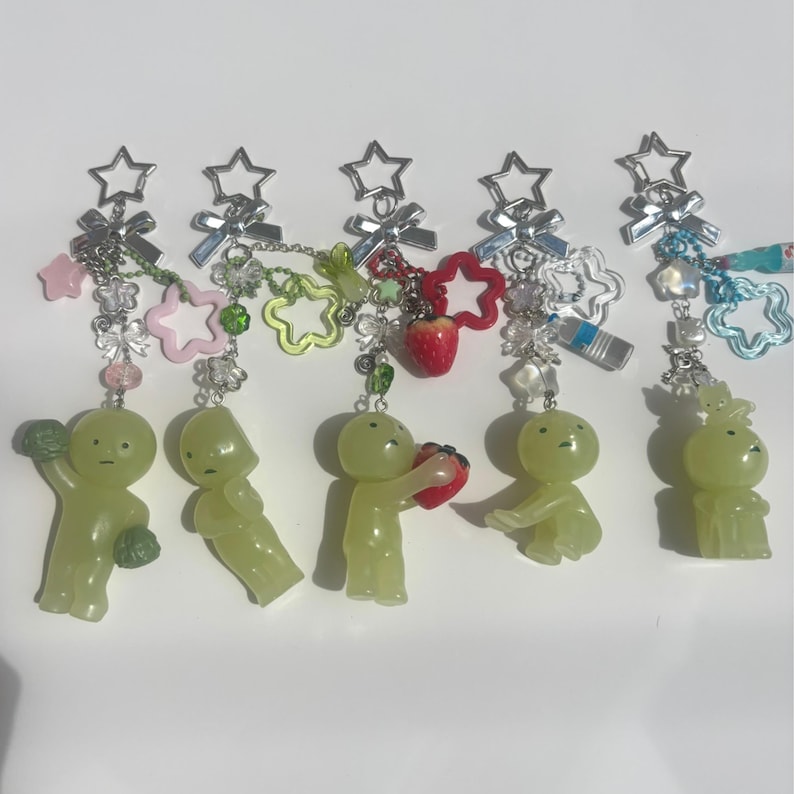 Peut inclure: Cinq porte-cl&eacute;s avec des figurines vertes, chacune tenant des objets diff&eacute;rents. Les porte-cl&eacute;s ont des fermoirs en forme d'&eacute;toile et sont orn&eacute;s de breloques, dont des &eacute;toiles, des fleurs et une fraise. Les figurines sont de couleur vert clair.