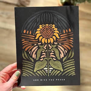 May include: A black and white print of a stylized sunflower with a colorful design. The print has the text "AND GIVE YOU PEACE" at the bottom.