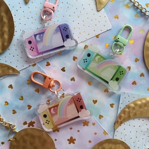 Peut inclure: Trois breloques colorées pour clés représentant une console Nintendo Switch stylisée avec un arc-en-ciel et un motif de lune. Les breloques sont en acrylique transparent et ont un anneau de clé en métal argenté.