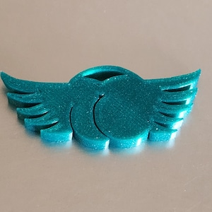 May include: A teal-colored, stylized wing emblem with a circular center and layered wing details. The emblem is made of a textured material and is set against a neutral background. The design is symmetrical and appears to be a decorative element.