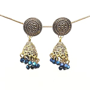 May include: A pair of gold-toned earrings with a circular top featuring a detailed pattern. The earrings have a bell-shaped bottom with intricate designs and dangling blue beads. The earrings are connected by a delicate gold chain.