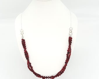 May include: A layered necklace with deep red beads. The necklace is displayed on a white mannequin. The beads are strung on a silver chain, with a silver clasp. The necklace is a statement piece.