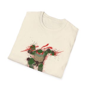 May include: Cream-colored t-shirt featuring a graphic of Raphael from Teenage Mutant Ninja Turtles. The design uses a splatter paint effect with red and green accents. The shirt is folded, showcasing the design prominently.