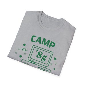Peut inclure: T-shirt gris chiné avec un motif vert « CAMP '85 ». Le motif comprend un ordinateur rétro, des étoiles et une planète. Le t-shirt est plié.