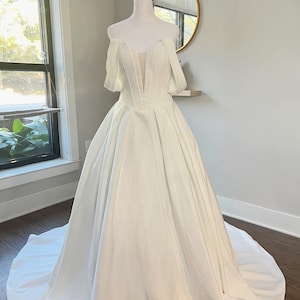 May include: A white off-the-shoulder wedding dress with a fitted bodice and a full skirt. The dress has a sweetheart neckline and a V-shaped cutout in the front.