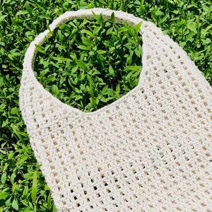 Può includere: Una borsa tote in crochet bianco con un'ampia apertura superiore. La borsa è realizzata con un semplice motivo a punto aperto e ha un aspetto naturale e rustico.