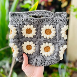Puede incluir: Una bolsa de mano de crochet gris con asa y un patr&oacute;n de margaritas de crochet blancas y marrones.