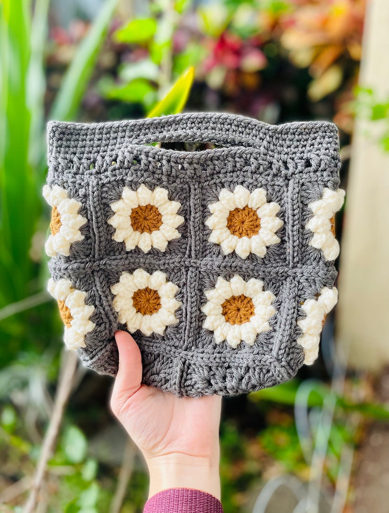 Puede incluir: Una bolsa de mano de crochet gris con asa y un patr&oacute;n de margaritas de crochet blancas y marrones.