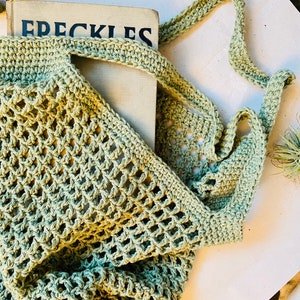 May include: A light green, open-weave crochet tote bag with a long strap. The bag is holding a book with the title "FRECKLES" visible. The bag is made of a soft, textured yarn.