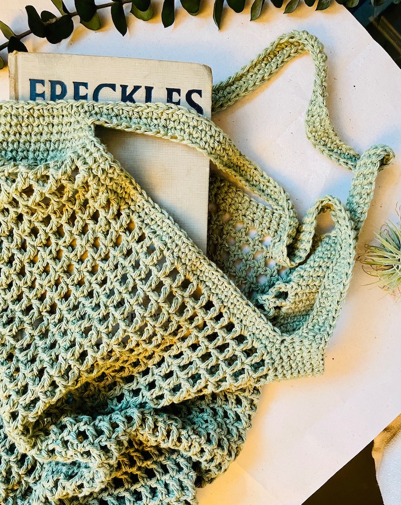 May include: A light green, open-weave crochet tote bag with a long strap. The bag is holding a book with the title "FRECKLES" visible. The bag is made of a soft, textured yarn.