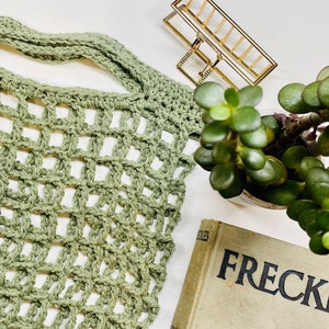 May include: A green crocheted market bag with a square grid pattern. The bag is lying on a white surface with a small potted plant and a gold metal hair clip.