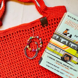 May include: A coral-colored crocheted tote bag with a long strap, a leather tag, and a book titled "Mosquitoes" by David Arnold. A beaded bracelet rests on the bag. The book cover features a bus illustration.