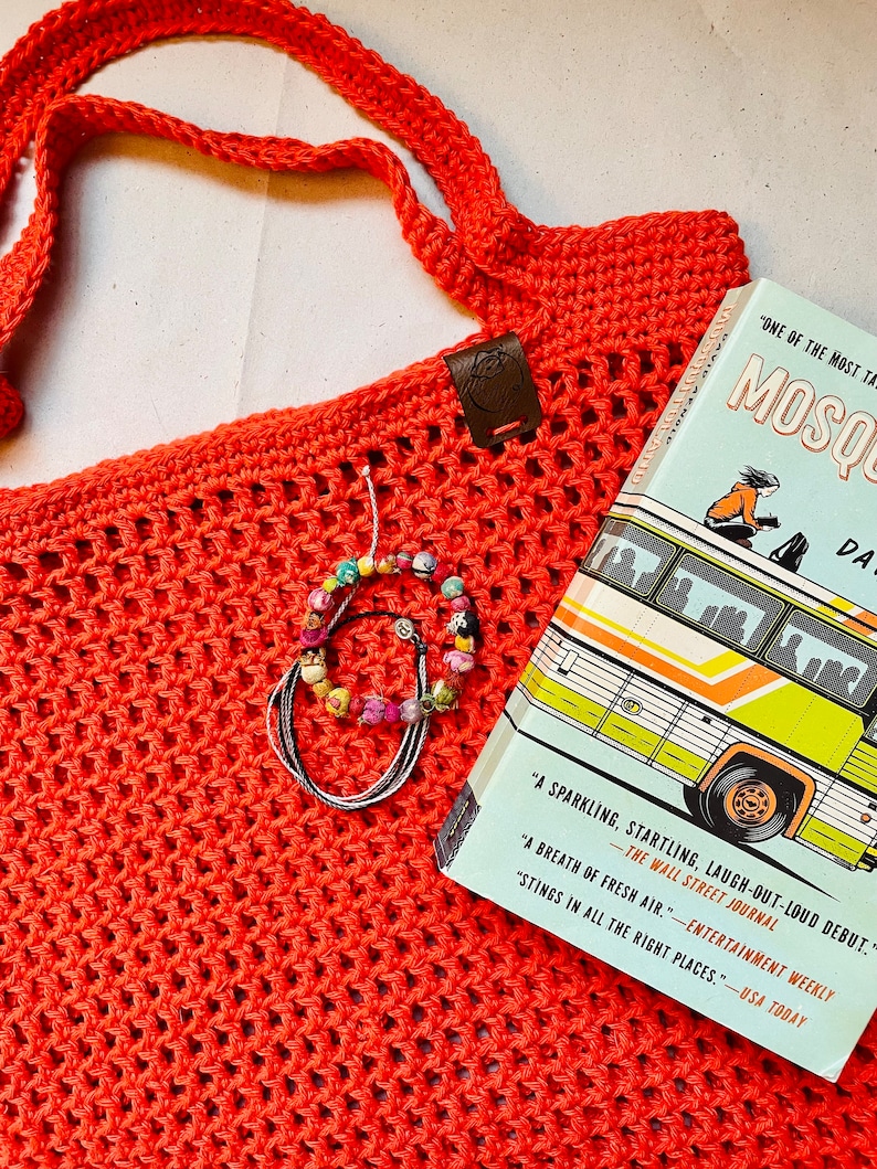 May include: A coral-colored crocheted tote bag with a long strap, a leather tag, and a book titled "Mosquitoes" by David Arnold. A beaded bracelet rests on the bag. The book cover features a bus illustration.