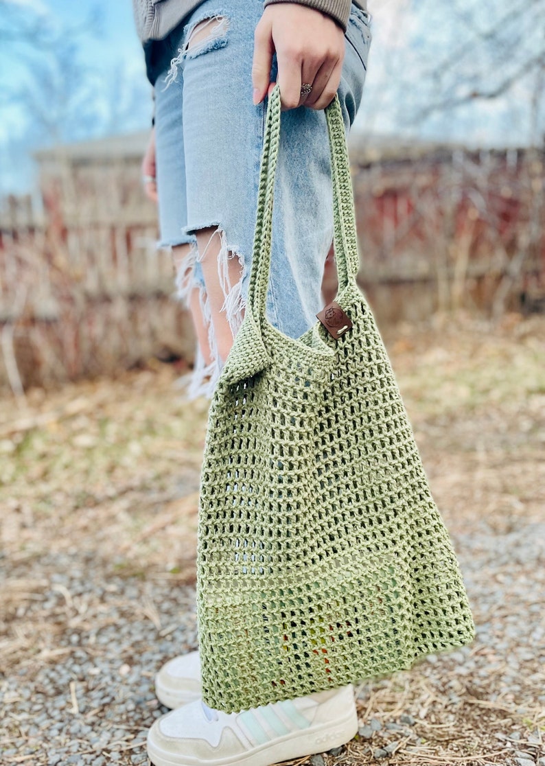 May include: A light green, open-weave crochet tote bag with a leather tag. The bag has a rectangular shape and long handles. The bag is being held outdoors, with a blurred background of a fence and foliage.