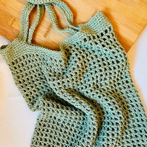 May include: A light green crocheted tote bag with long straps, resting on a wooden cutting board. The bag has an open, net-like weave. The straps are knotted at the top. The bag is designed for carrying groceries or other items.
