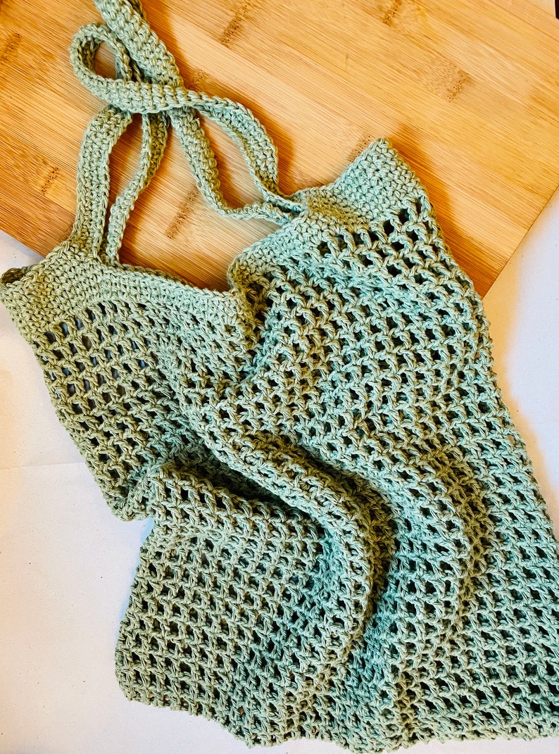 May include: A light green crocheted tote bag with long straps, resting on a wooden cutting board. The bag has an open, net-like weave. The straps are knotted at the top. The bag is designed for carrying groceries or other items.
