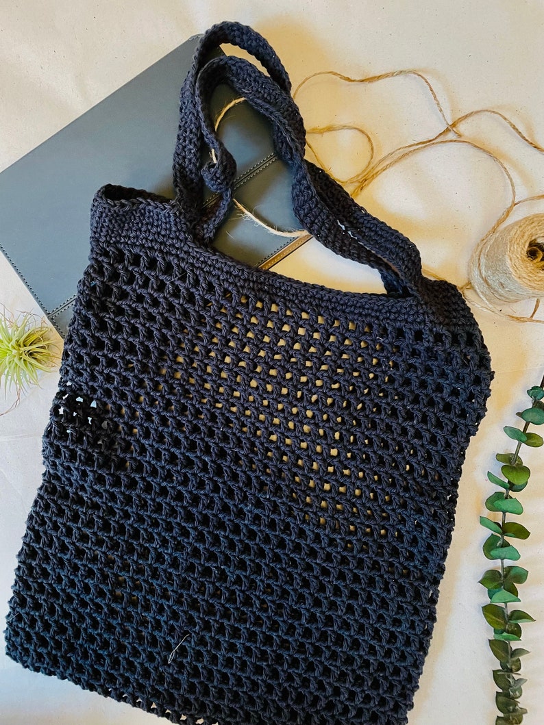 May include: A navy blue crocheted tote bag with a net-like design. The bag has two handles and is sitting on a light-colored surface. A gray notebook and a ball of twine are also visible.