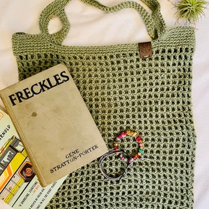 May include: A sage green crocheted tote bag with a rectangular shape and open weave design. The bag has two handles and a small leather tag. A vintage book titled "FRECKLES" and a colorful beaded bracelet are inside the bag.