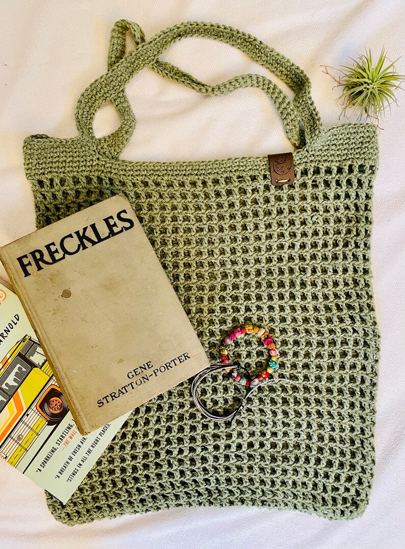May include: A sage green crocheted tote bag with a rectangular shape and open weave design. The bag has two handles and a small leather tag. A vintage book titled "FRECKLES" and a colorful beaded bracelet are inside the bag.
