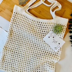 May include: Cream-colored, open-weave crochet tote bag with sturdy handles. A small tag reads "HANDMADE WITH LOVE." Another tag says "thank you." The bag is displayed on a wooden cutting board.