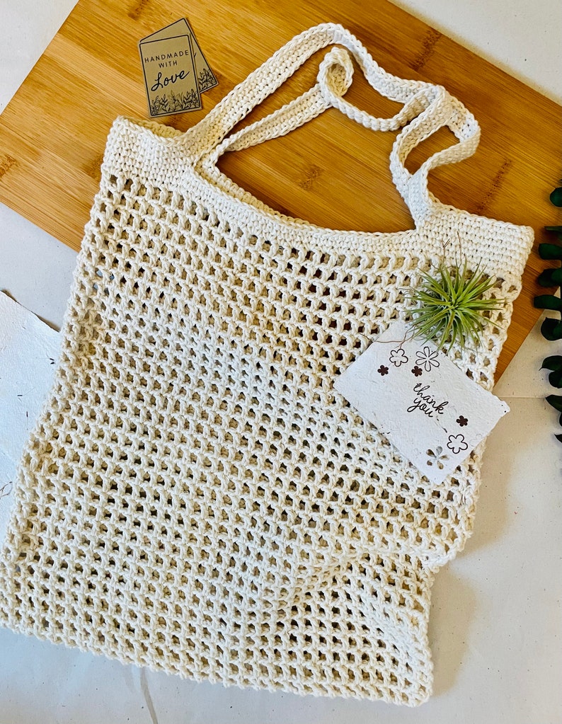 May include: Cream-colored, open-weave crochet tote bag with sturdy handles. A small tag reads "HANDMADE WITH LOVE." Another tag says "thank you." The bag is displayed on a wooden cutting board.