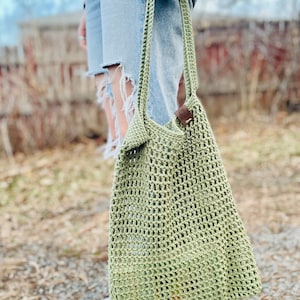 May include: A green crocheted tote bag with a brown leather strap. The bag is being held by a person wearing ripped jeans and white sneakers.