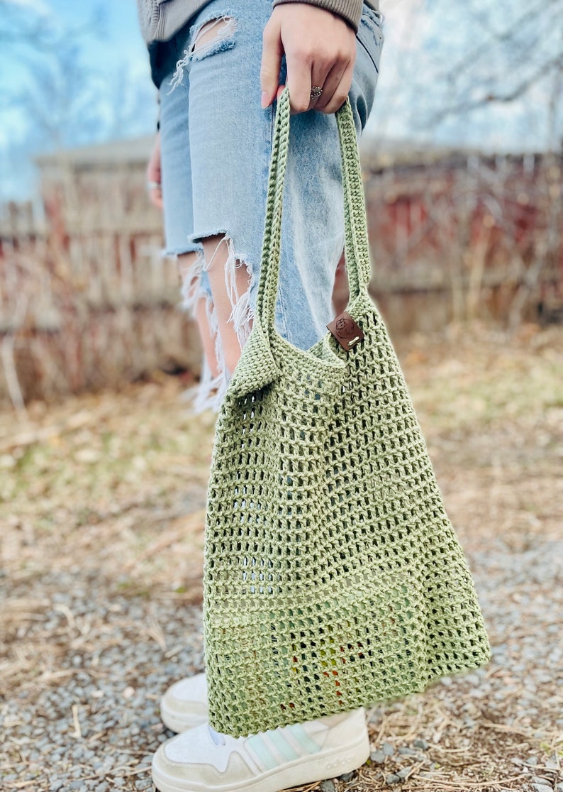 May include: A green crocheted tote bag with a brown leather strap. The bag is being held by a person wearing ripped jeans and white sneakers.