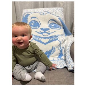 May include: A baby sitting on a couch with a blue and white knitted blanket featuring a cartoon dog design. The blanket has a white background with a blue dog face and a blue collar.