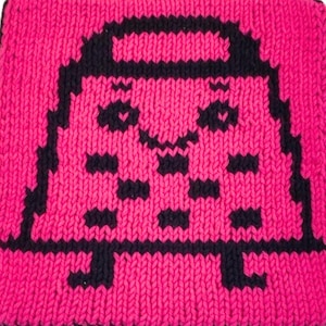 May include: A square, knitted item in bright pink with a black pixelated design of a smiling face and a rectangular body. The edges are finished with a black border. The item is likely a decorative piece or a small accessory.