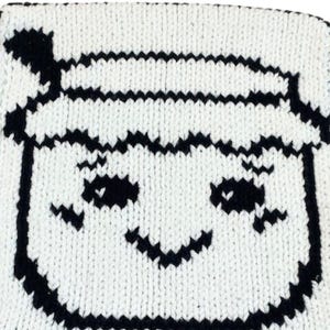 May include: A square, knitted item featuring a smiling face design. The face is outlined in black against a white background. The design includes a hat, eyes, and a curved mouth, all in black.