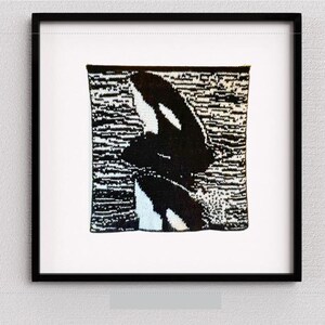 Puede incluir: Obra de arte enmarcada con un diseño de orca tejida en blanco y negro. La orca se representa sobre un fondo texturizado, enmarcada en un marco cuadrado negro. La obra está montada sobre un fondo blanco.