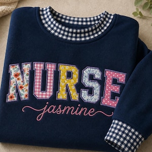 May include: A navy blue long-sleeve sweatshirt featuring the word NURSE in large, multi-patterned fabric letters and the name jasmine embroidered in pink script below. The collar and sleeve cuffs have a navy and white gingham check pattern.