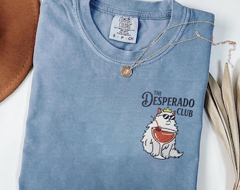 Könnte beinhalten: Hellblaues T-Shirt mit dem Text "The Desperado Club" und einer Cartoon-Katze mit Sonnenbrille und Krone, die einen Cocktail hält. Eine goldene Halskette und ein brauner Hut sind ebenfalls zu sehen.